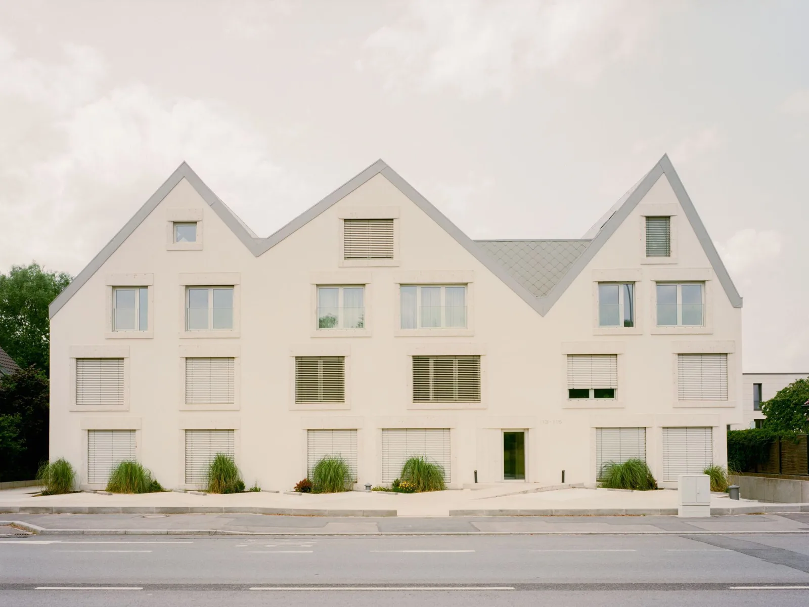 White residential building with three steep gables and green shuttered windows beside a road