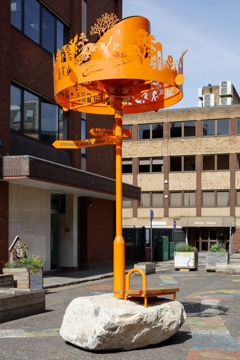 Orange sculptural lamppost with cut-out figures on top, standing on a large boulder in a paved street