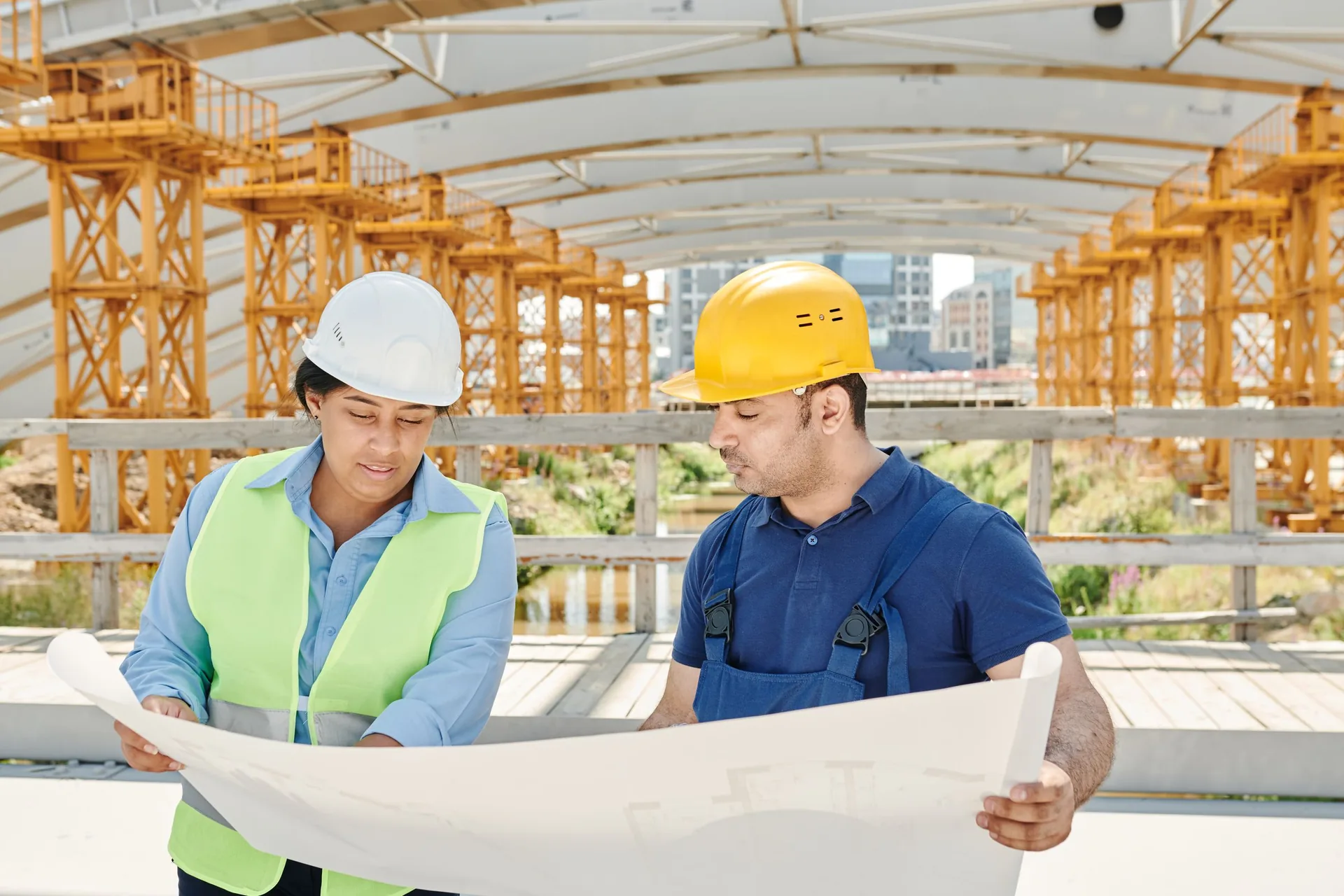 Two people in hard hats, one in a high-visibility vest, reading a large drawing together on a construction site