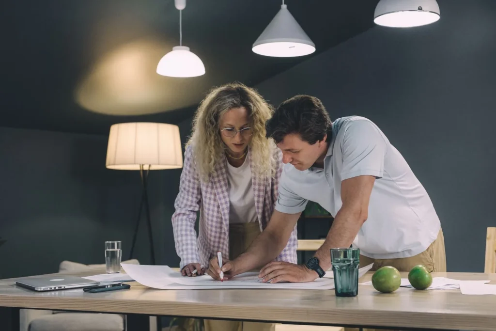 Two designers marking up a large printed drawing together on a table under pendant lights