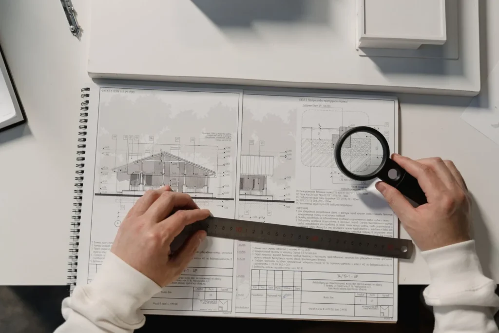 Hands holding a steel ruler and a magnifying glass over a bound set of house drawings and specifications