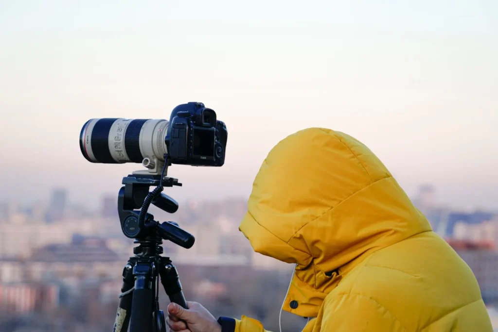 Photographer in a yellow hooded jacket beside a long-lens camera on a tripod above a hazy city, the patient setup behind architectural photography