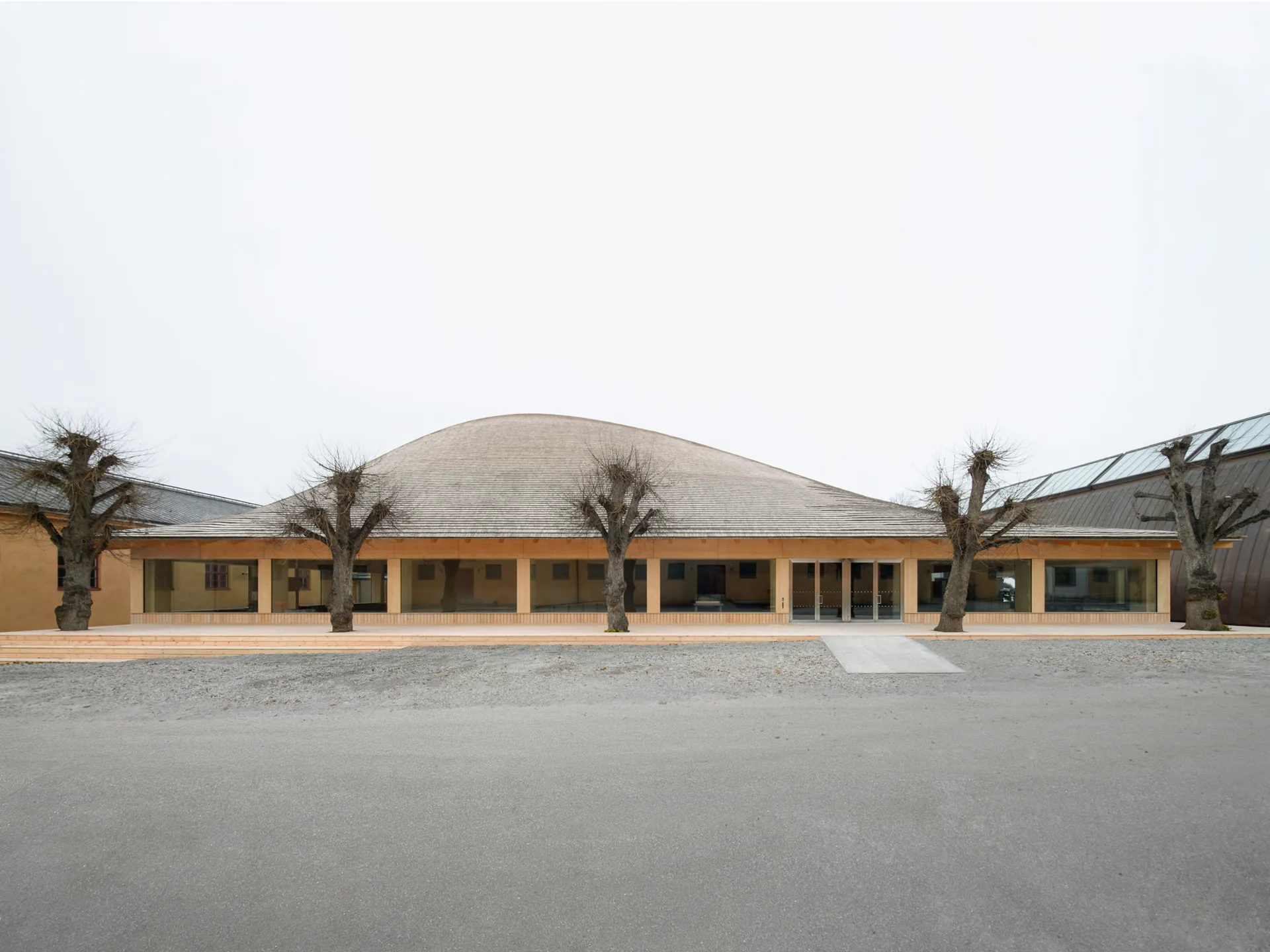 Low glazed facade under the curved shingled roof with pollarded trees in front, Wisdome Stockholm by Elding Oscarson