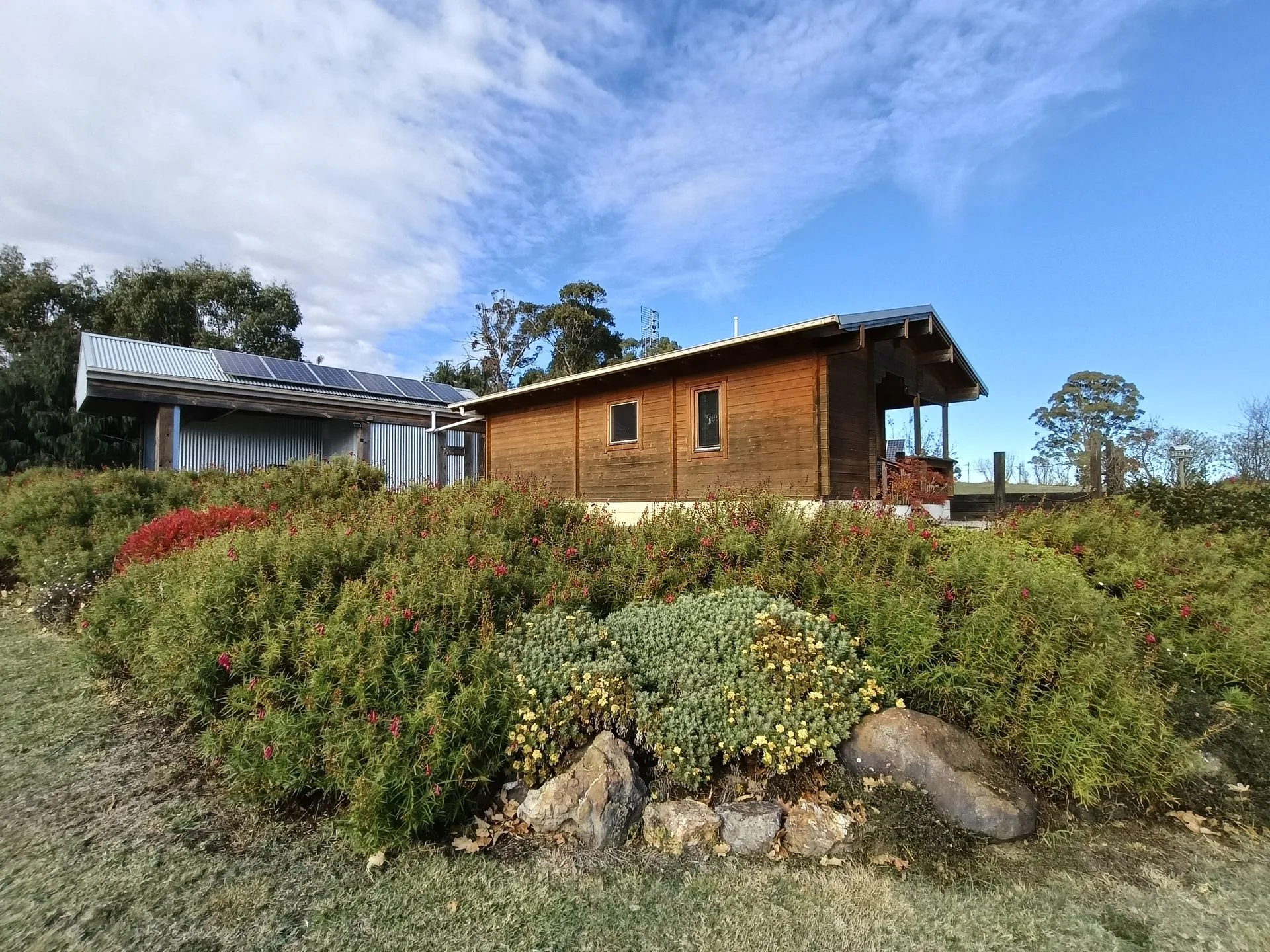 Small wooden cabin with a solar array on an adjoining metal-roofed outbuilding, surrounded by flowering shrubs
