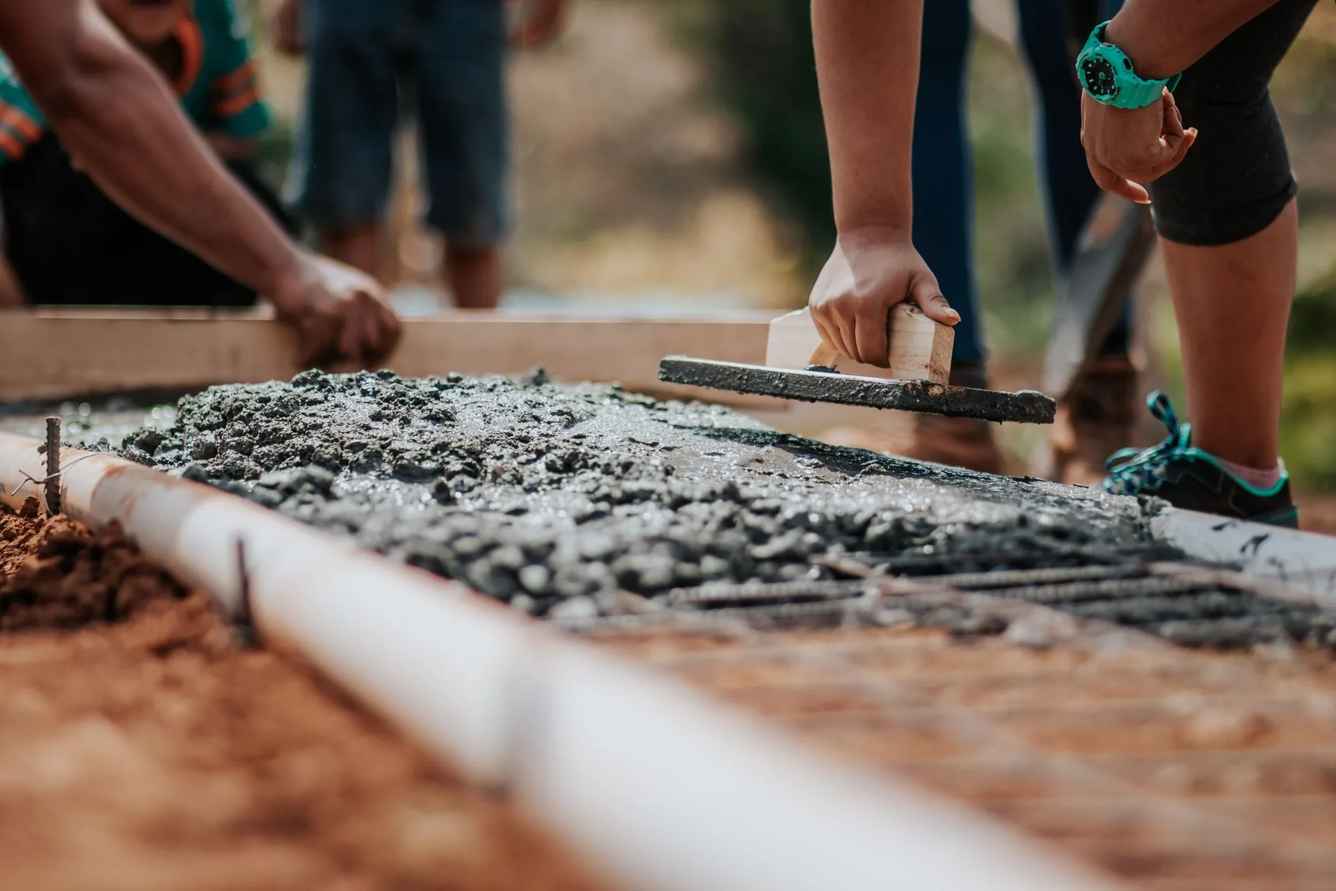 Workers leveling freshly poured concrete over steel mesh with a timber screed board beside a pipe