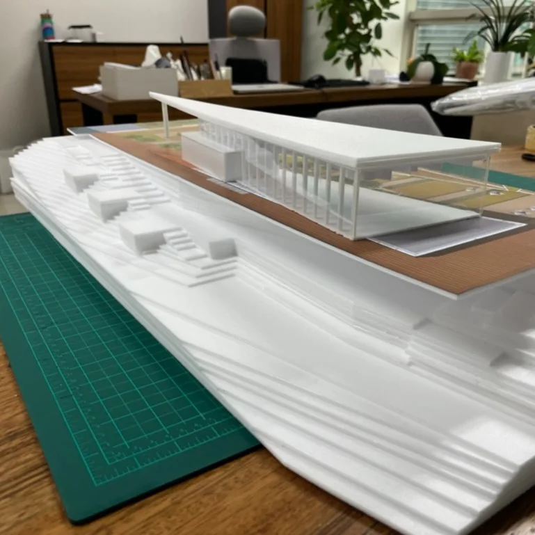 Desk photo of a white house model with a canopy roof and stepped terrain beside a cutting mat