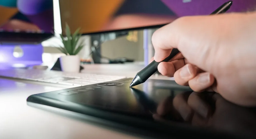 Hand using a stylus on a graphics tablet at a desk in front of a monitor