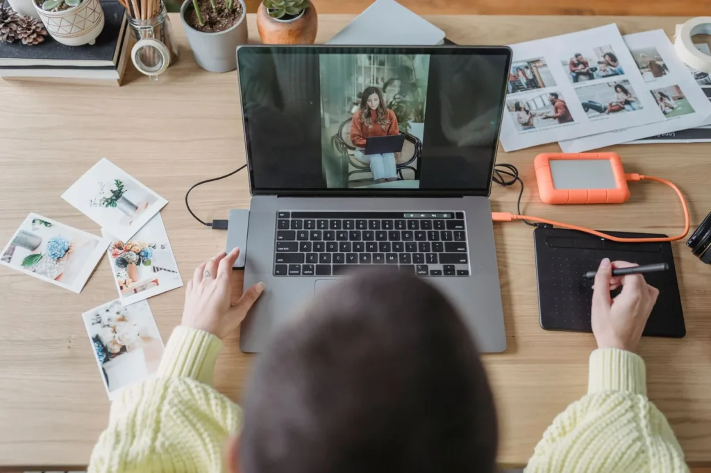 Designer retouching an image on a laptop surrounded by printed photographs and a tablet