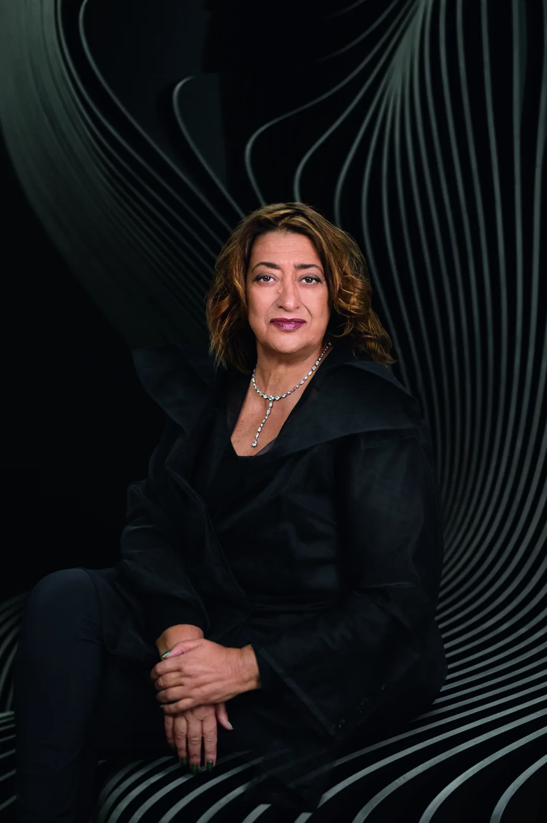 Portrait of Zaha Hadid dressed in black, seated against a dark backdrop of curving striped lines