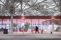 Glass front of the red waiting area under the white steel roof structure, with a passenger walking past benches, Zvonařka Bus Station by CH+K